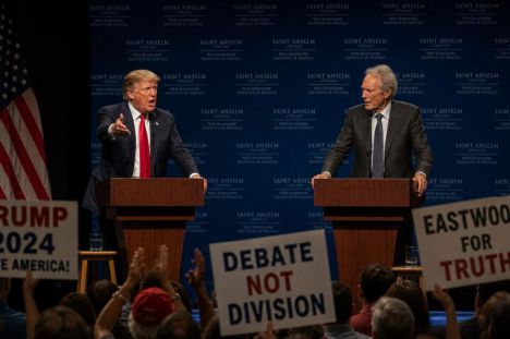 Trump y Clint Eastwood debaten en un auditorio, mientras la multitud aplaude y protesta con pancartas.