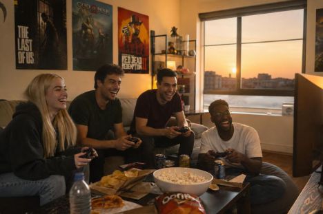 Grupo de amigos disfruta de videojuegos en sala de estar iluminada por el atardecer, con snacks y decoración gamer.