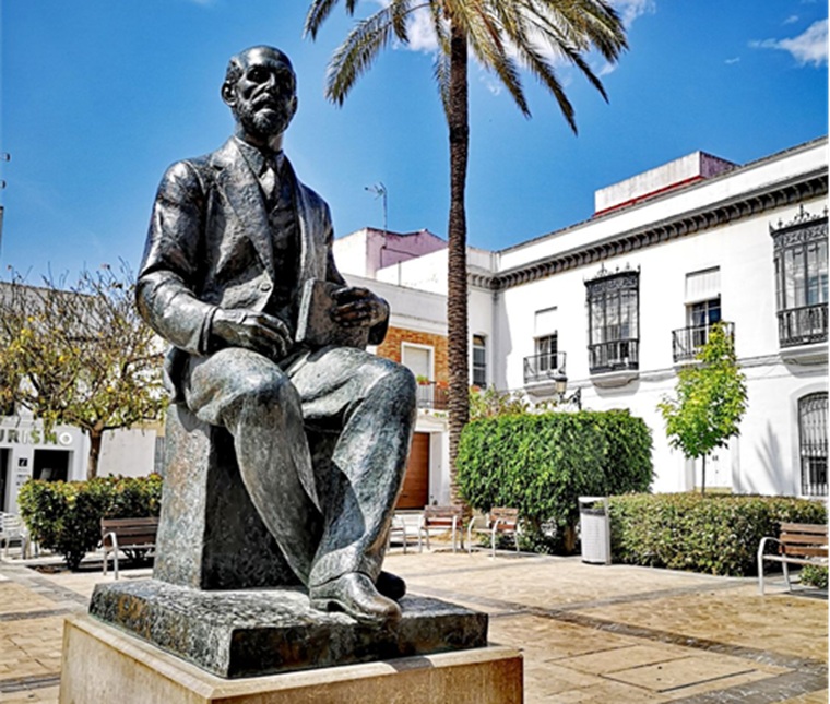 Estatua de granito que representa al genio lírico moguereño en su plenitud creativa, situada en la plaza del Cabildo, frente a la fachada del edificio del Ayuntamiento. Es obra del gran escultor valenciano Octavio Vicent que la realizó en 1981 con motivo del centenario del nacimiento del poeta. En la actualidad está rodeada de una fuente y junto a ella se celebran numerosos actos literarios. (Ayuntamiento de Moguer)